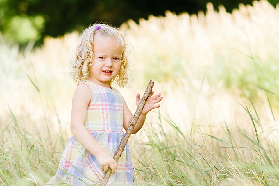 Toddler girl in plaid dress, walking through tall grass with a stick during a summer family photo session at Lee Martinez Park.