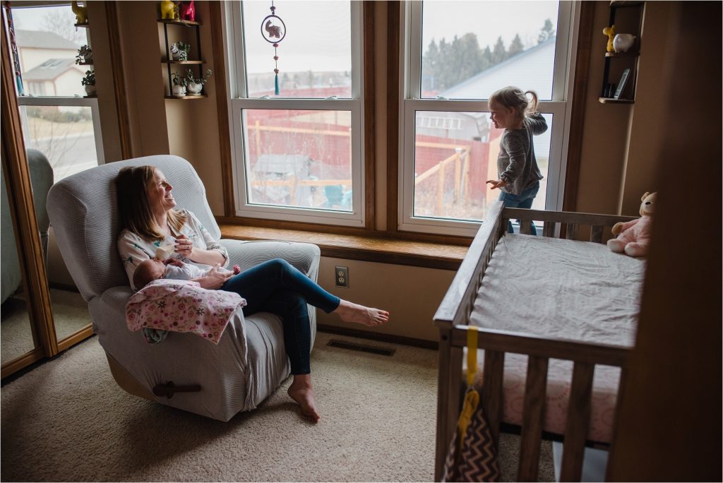 Mom sitting in chair feeding baby while laughing with toddler.