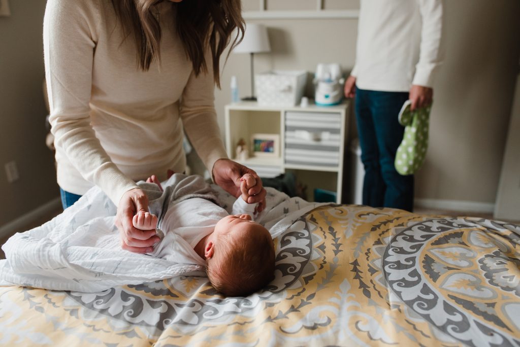 Lifestyle photography photo of mom unswaddling baby on bed while dad warms bottle in background.