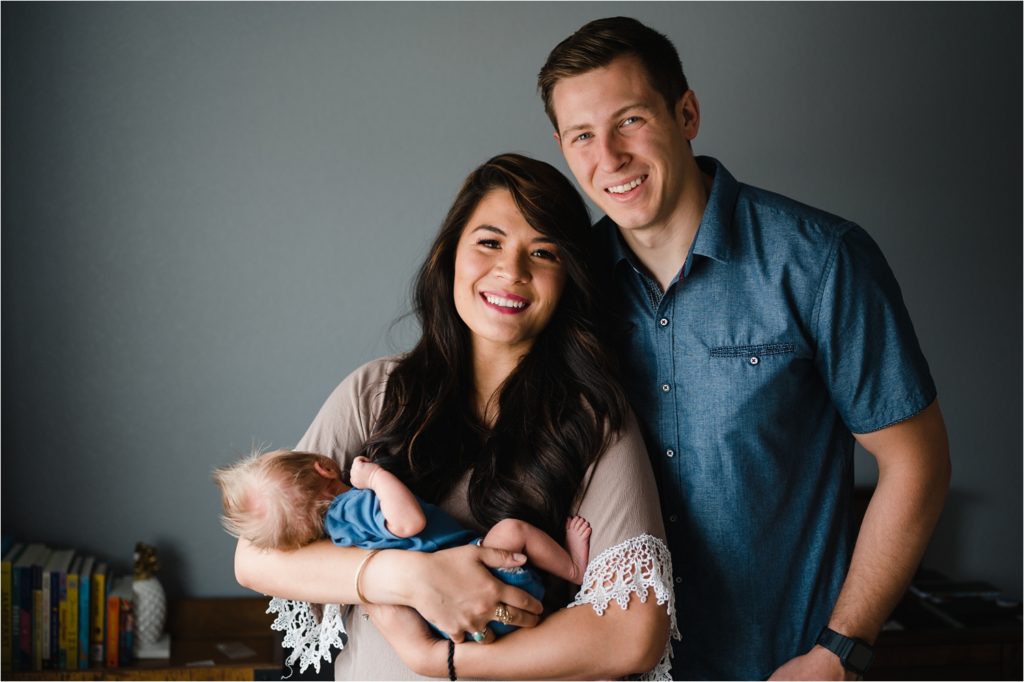 Parents smiling at camera while holding baby during in home newborn photography session.