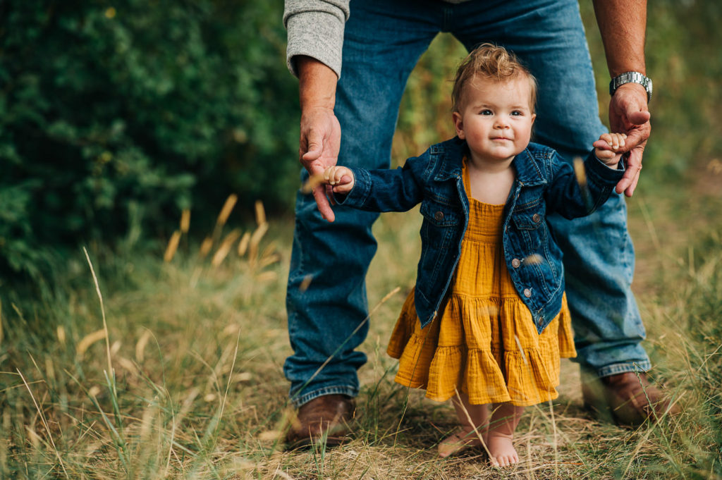 Baby in yellow dress and jean jacket holding dad's fingers while learning to walk during family photo session.
