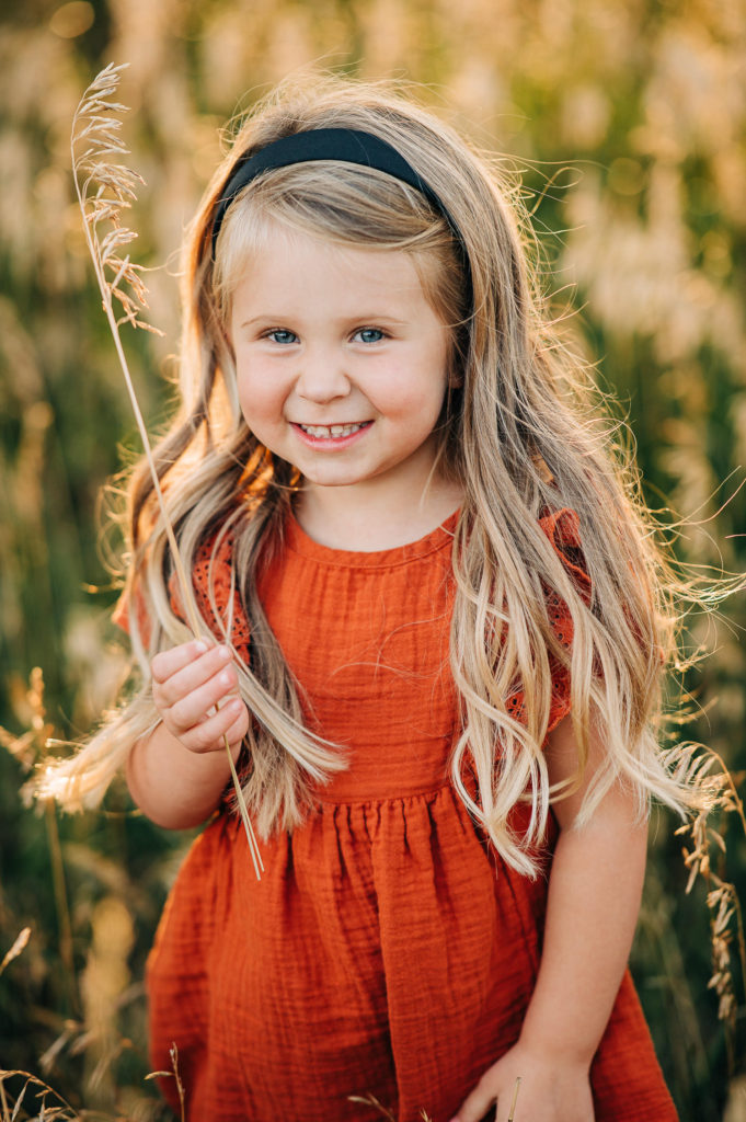 Girl in orange dress is smiling and laughing while holding stalk of grass.