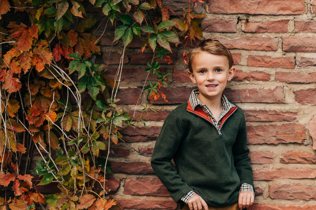 Boy in green sweater and khaki pants is laughing while leaning on wall.