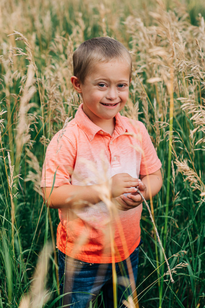 Boy in orange shirt in tall grass, smiling at camera.