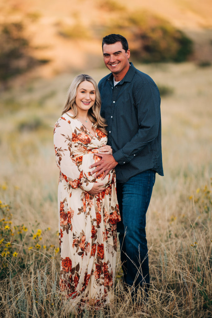Couple stands in grass field while smiling at the camera.