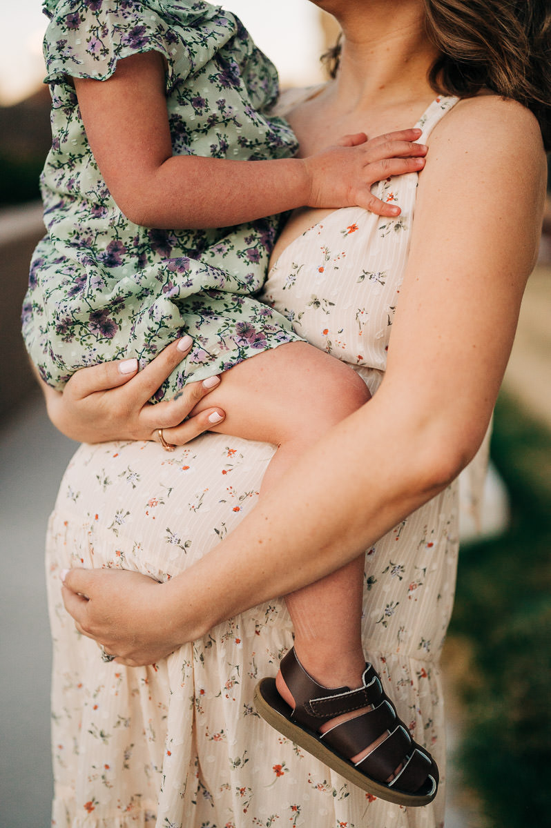 Mom to be in sundress, holding 3 year old in flowered dress on her belly.