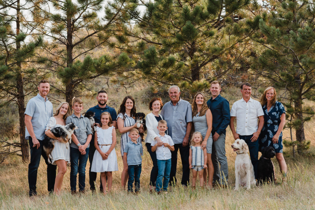 Large family in white and blue and gray outfits, standing in tall grass in front of pine trees in Wyoming.