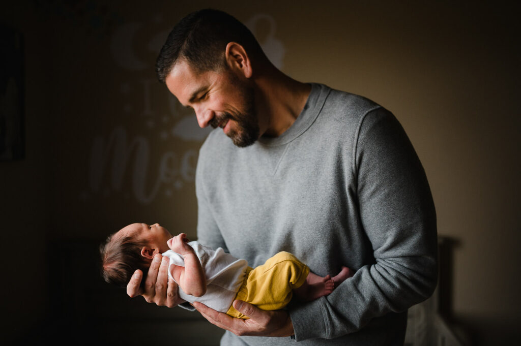 Dad in gray sweater holding newborn baby girl in yellow pants.