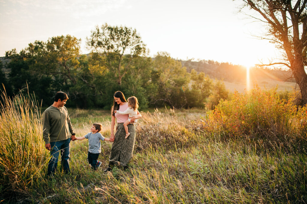 Sun sets behind family holding hands, while standing in field of tall grass.