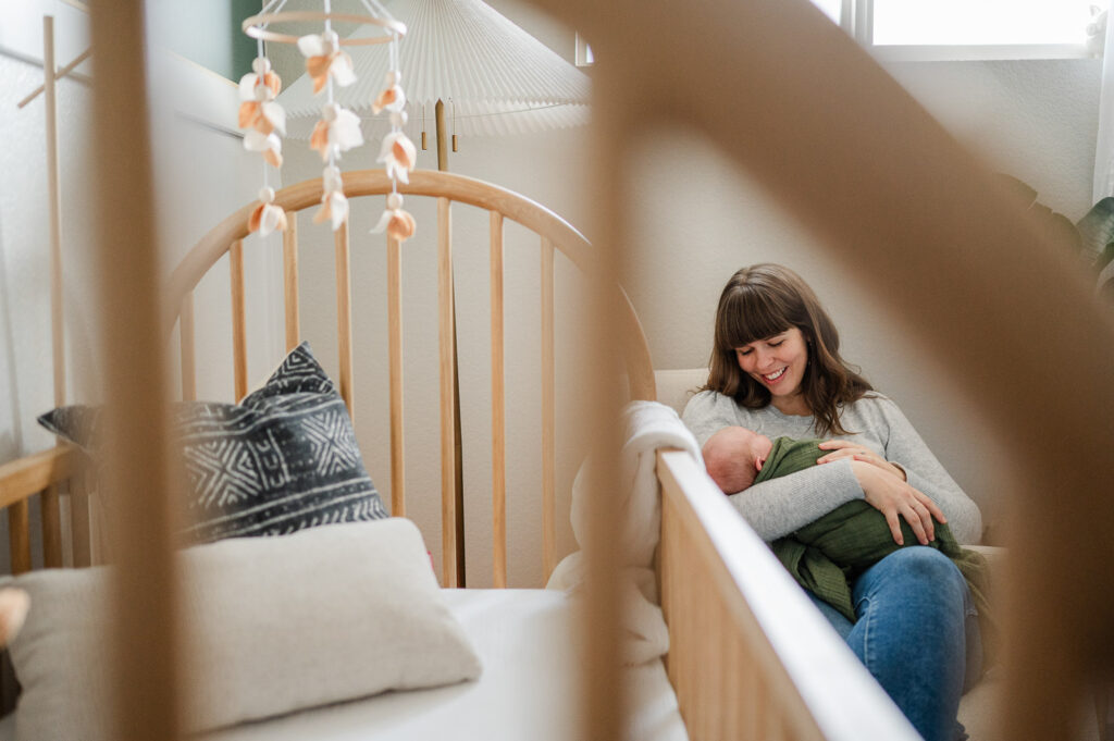New mom sits in chair next to crib, and smiles at her baby.