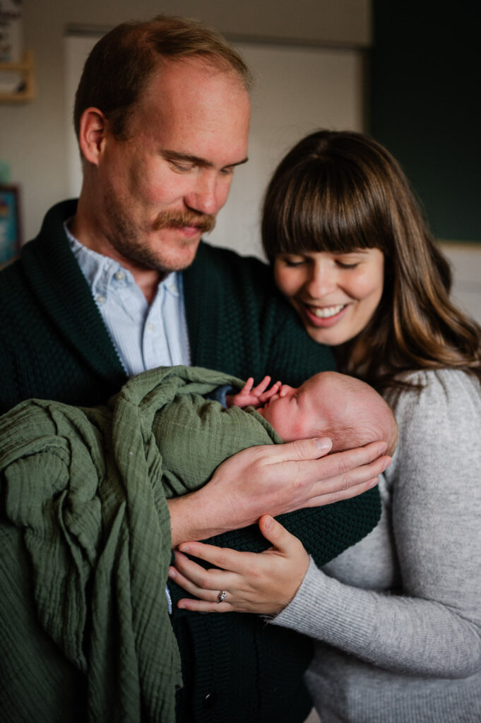 Newborn baby is wrapped in a green swaddle while being snuggled by his parents during a Denver newborn photoshoot.