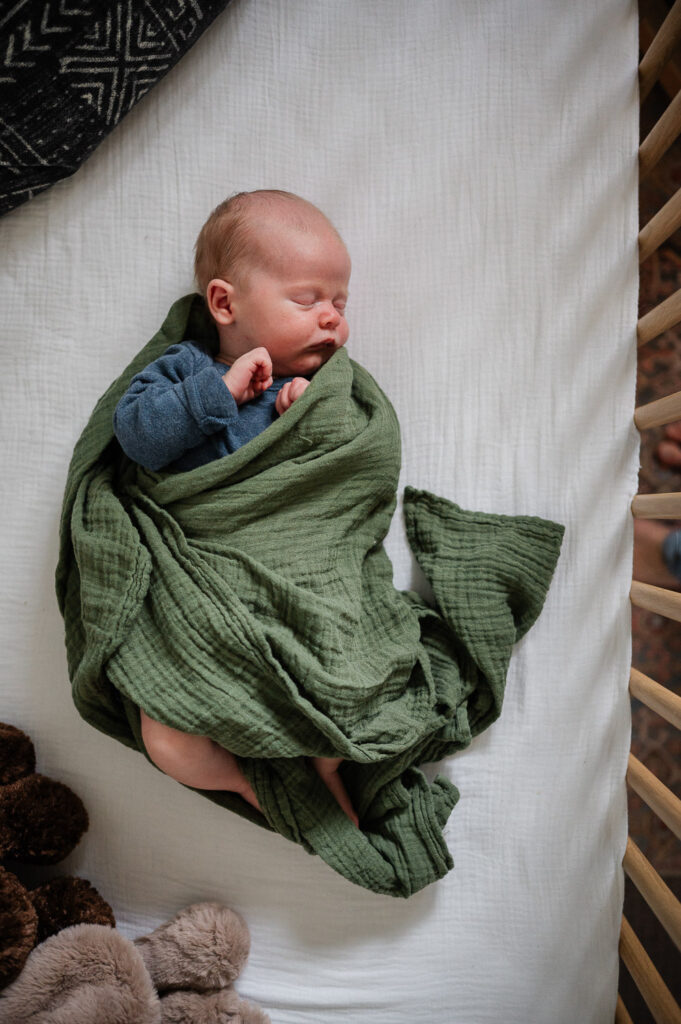 Newborn baby in blue onesie is wrapped up in a green blanket while sleeping in his crib.